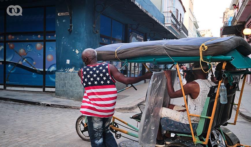 Hombre cubano con camiseta de la bandera estadounidense empuja un bicitaxi que trasporta a otras personas, La Habana. Cuba.