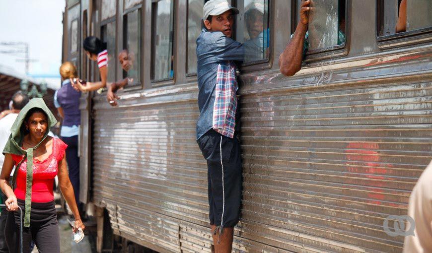Cuba. Estación de ferrocarril. Una persona se sostiene de una ventana. Otros transeúntes caminan.