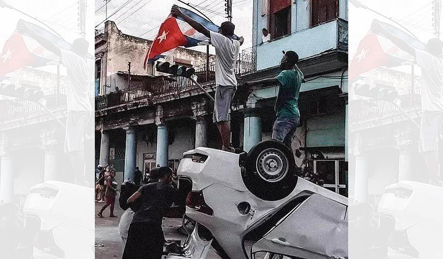 Una de las imágenes icónicas de las protestas del 11J muestra una patrulla policial volcada y encima dos jóvenes, uno de ellos ondea una bandera cubana ensangrentada